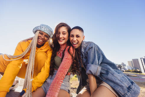 Generation Z Freunde, die eine gute Zeit im Freien haben. Drei glückliche Freunde, die in die Kamera lächeln, während sie auf einer Mauer in der Stadt sitzen. Fröhliche weibliche Jugendliche, die die gemeinsame Zeit genießen. - JLPSF06852