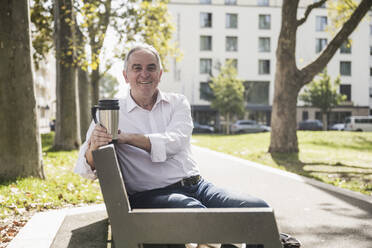 Smiling senior man holding insulated drink container sitting on bench - UUF27513