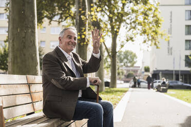 Smiling senior man with walking cane waving and sitting on bench - UUF27528