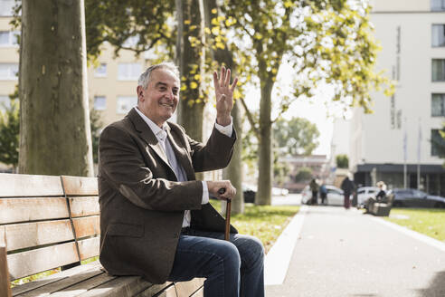 Lächelnder älterer Mann mit Gehstock, der auf einer Bank sitzt und winkt - UUF27528