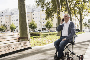 Lächelnder älterer Mann, der auf einer Gehhilfe sitzend mit einem Mobiltelefon spricht - UUF27533