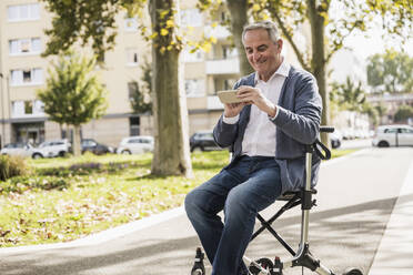 Glücklicher älterer Mann, der ein Smartphone benutzt und auf einer Gehhilfe sitzt - UUF27536