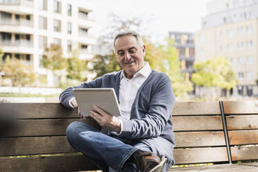 Lächelnder älterer Mann mit Tablet-PC auf einer Bank sitzend - UUF27542