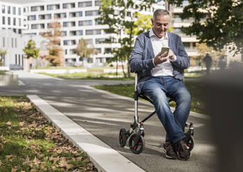 Lächelnder älterer Mann, der ein Smartphone benutzt und auf einer Gehhilfe sitzt - UUF27549