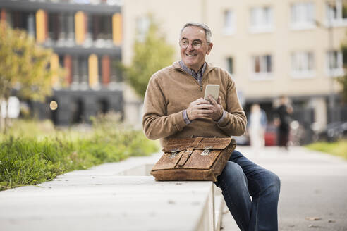 Happy senior man holding mobile phone sitting on concrete bench - UUF27573