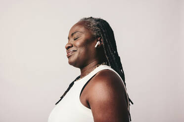 Middle-aged woman with dreadlocks listening to music using wireless earbuds. Mature woman enjoying her favourite playlist while standing against a studio background. - JLPSF06883