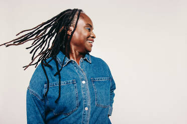 Dreadlocks fun. Excited mature woman smiling and whipping her dreadlocks while standing against a grey background. Happy middle-aged woman wearing a denim jacket and make-up in a studio. - JLPSF06893