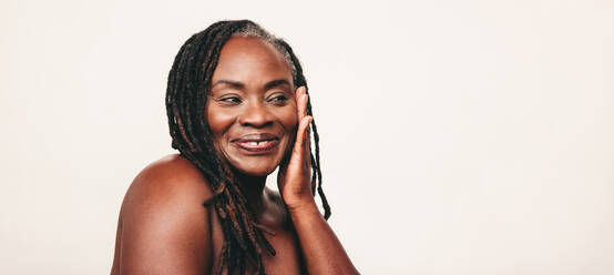 Happy mature woman with dreadlocks smiling while touching her flawless skin. Cheerful dark-skinned woman embracing her beautiful melanated skin. Mature black woman ageing gracefully. - JLPSF06896