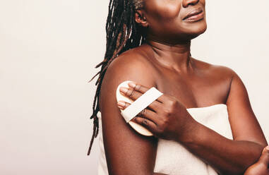 Mature woman exfoliating her skin with a brush against a studio background. Woman with dreadlocks dry brushing her arm while wrapped in a bath towel. Middle-aged woman taking care of her ageing body. - JLPSF06937