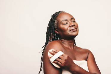 Woman exfoliating her skin with a brush while standing against a studio background. Woman with dreadlocks dry brushing her arm while wrapped in a bath towel. Mature woman taking care of her ageing body. - JLPSF06939