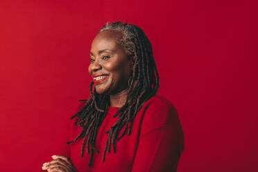 Cheerful black woman smiling and looking away while standing against a red background. Happy mature woman embracing her natural hair with confidence. - JLPSF06946