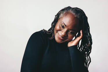 Confident mature woman smiling at the camera while touching her dreadlocks. Happy middle-aged woman embracing her natural hair with pride. Cheerful black woman standing against a grey background. - JLPSF06973