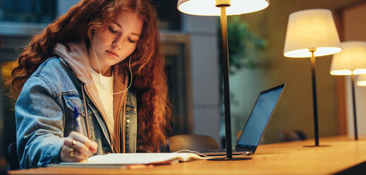 Mädchen macht sich Notizen in ihrem Buch unter der Lampe in der Bibliothek, mit Laptop auf dem Schreibtisch. Junge Frau in der College-Bibliothek, die ihre Aufgabe erledigt. - JLPSF06982