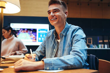 Glücklicher junger Mann, der in der Bibliothek sitzt, wegschaut und lächelt, während er studiert. Student, der in der Universitätsbibliothek studiert. - JLPSF06987