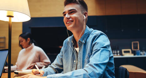 Lächelnder männlicher Student mit Kopfhörern, der in der Universitätsbibliothek lernt. Glücklicher junger Mann in der Universität, der wegschaut und lächelt. - JLPSF06988