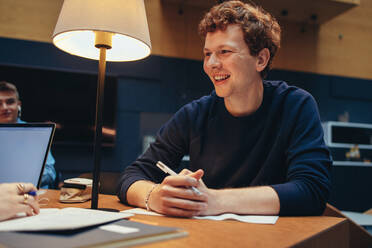 Junger Mann lächelnd, während er in der Bibliothek studiert. Student, der glücklich aussieht, während er in der College-Bibliothek studiert. - JLPSF06992