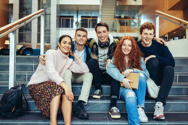 Porträt von Studenten, die auf einer Treppe sitzen und in die Kamera lächeln. Glückliche High-School-Studenten, die auf dem Campus sitzen. - JLPSF07001