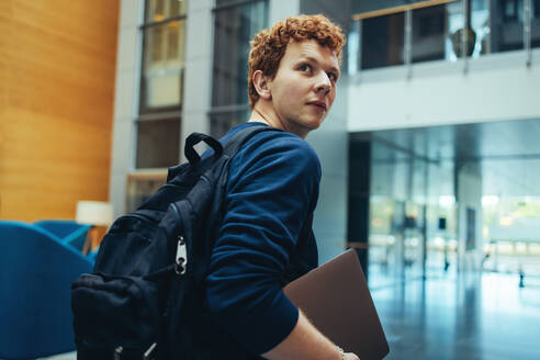 College-Student trägt seine Tasche und Laptop auf dem Campus. Junger Mann dreht sich über die Schulter zurück und geht auf dem College-Campus. - JLPSF07008