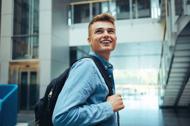 Junger Student mit Tasche lächelnd in High-School-Campus. Glücklicher junger Mann schaut weg und lächelt nach dem Unterricht. - JLPSF07011