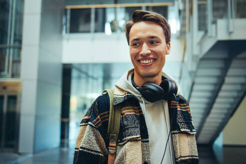 Männlicher Student, der wegschaut und auf dem Campus lächelt. Glücklicher junger Mann in Freizeitkleidung und Kopfhörern in der High School. - JLPSF07013