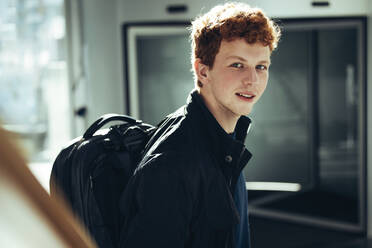 Junger Mann mit Tasche auf dem Weg zum Unterricht in der High School. Männlicher Schüler auf dem Weg zum Unterricht und Blick in die Kamera. - JLPSF07023