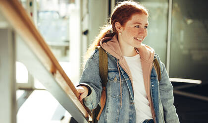 Glückliche junge Studentin auf dem Weg zum Unterricht, die auf einer Treppe steht. Ein Mädchen, das auf einer Treppe steht, schaut weg und lächelt auf dem Campus. - JLPSF07024