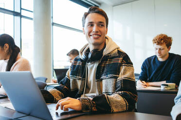 Junger Mann sitzt am Schreibtisch im Klassenzimmer, schaut weg und lächelt. Student mit Laptop auf dem Schreibtisch im Unterricht. - JLPSF07028