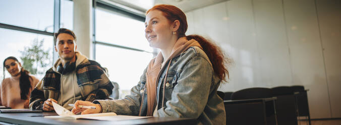 Junge Schülerin in einem Klassenzimmer mit ihren Freunden, die herüberschauen. Schülerin beantwortet Frage in einem Klassenzimmer. - JLPSF07032