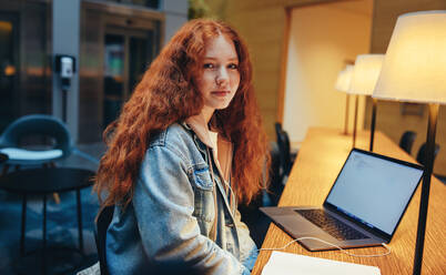 Porträt eines Mädchens mit gefärbten roten Haaren, das in die Kamera schaut, während es am Bibliothekstisch sitzt. Junge Frau in der Bibliothek bei Nacht. - JLPSF07042