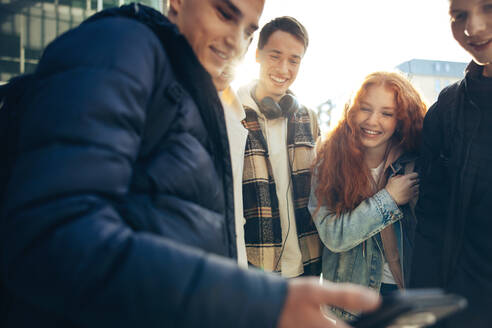 Ein junger Mann zeigt seinen Freunden auf dem College-Campus sein Handy, während sie etwas auf dem Handy anschauen. - JLPSF07050