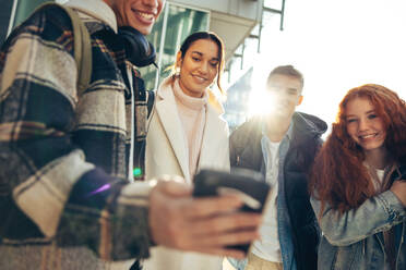 Freunde im Freien mit Blick auf Handy mit Sonne scheint. Glückliche Männer und Frauen lächeln zusammen beim Blick auf Handy. - JLPSF07051