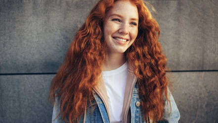 Beautiful young woman against wall. Student with red dyed hair looking away and smiling - JLPSF07058