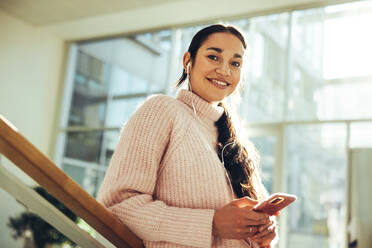 Mädchen in der Universität schaut mit Handy in der Hand in die Kamera. Fröhliche junge Frau mit Handy auf dem College-Campus. - JLPSF07096