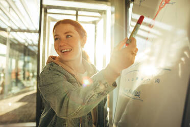 Studentin zeigt auf Whiteboard und lächelt. Glückliche junge Frau zeigt Whiteboard in der Universität. - JLPSF07105