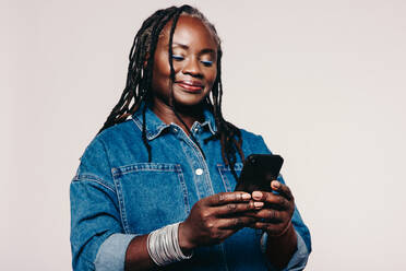 Mature woman reading a text message on a smartphone while standing against a grey background. Stylish woman with dreadlocks wearing a denim jacket and make-up in a studio. - JLPSF07131