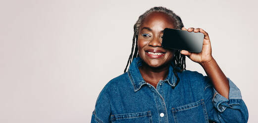 Cheerful woman looking away with a smile while holding a smartphone in front of her eye. Happy mature woman with dreadlocks wearing a denim jacket and make-up in a studio. - JLPSF07133