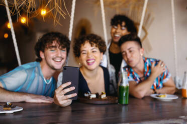 Eine Gruppe von Freunden macht ein gemeinsames Selfie in einem Restaurant. Vier junge queere Menschen lächeln fröhlich, während sie den Moment festhalten. Freunde sitzen beim Mittagessen zusammen und kommen sich näher. - JLPSF07143