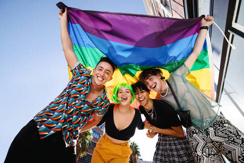 Four young people celebrating their queer identities at a gay pride parade. Group of young LGBTQ+ people smiling cheerfully while raising the rainbow pride flag. Friends standing together outdoors. - JLPSF07155