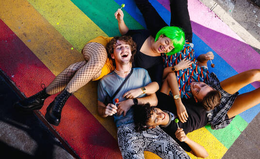 Four young queer people lying down on rainbow colours together. Group of happy LGBTQ+ people smiling cheerfully while each holding a lollipop. Friends bonding together outdoors. - JLPSF07160