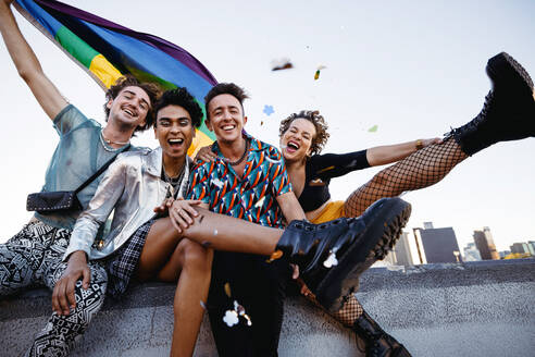 Friends celebrating gay pride while sitting together. Four members of the LGBTQ+ community smiling cheerfully while raising the pride flag. Group of queer individuals celebrating together outdoors. - JLPSF07177