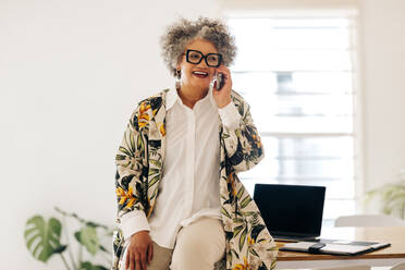 Cheerful businesswoman smiling happily while speaking with her business partners on a phone call. Mature businesswoman making business plans while sitting in a modern office. - JLPSF07192