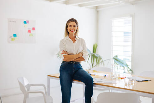 Ein Manager lächelt in die Kamera, während er auf einem Konferenztisch in einem modernen Sitzungssaal sitzt. Eine fröhliche Geschäftsfrau wartet darauf, eine Sitzung in einem kreativen Büro zu beginnen. - JLPSF07230