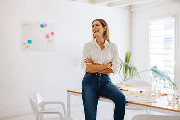 Glückliche Geschäftsfrau, die mit einem Lächeln wegschaut, während sie an einem Konferenztisch in einem modernen Sitzungssaal sitzt. Erfolgreiche Geschäftsfrau, die in einem kreativen Büro arbeitet. - JLPSF07233