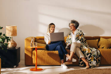 Zwei glückliche Geschäftsfrauen, die in einer Bürohalle gemeinsam an einem Laptop arbeiten. Erfolgreiche Unternehmerinnen, die in einem von Frauen geführten Betrieb auf einer Couch sitzen. - JLPSF07266