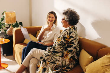 Happy female entrepreneurs laughing cheerfully while working on a laptop in an office lobby. Two successful businesswomen sitting on a couch in a woman-owned workplace. - JLPSF07268