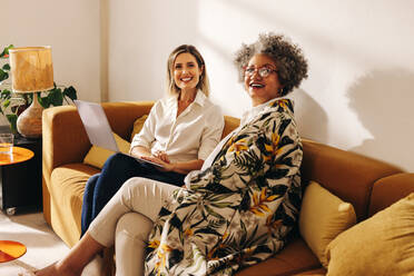 Cheerful businesswomen smiling happily while sitting on a couch in a modern office. Two successful businesswomen working in the lobby of a woman-owned workplace. - JLPSF07269