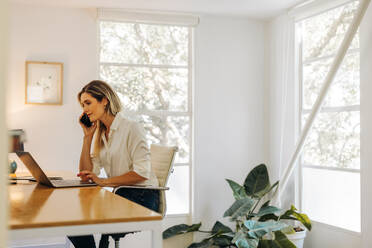 Junge Geschäftsfrau beim Telefonieren, während sie in ihrem Heimbüro am Laptop arbeitet. Kreative Geschäftsfrau, die Pläne mit ihren Geschäftskunden schmiedet. - JLPSF07283