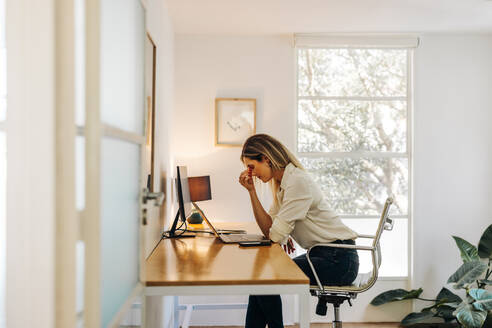 Exhausted businesswoman having a headache in her office. Overworked businesswoman experiencing work fatigue while siting in front of a laptop. - JLPSF07286