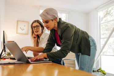 Two young businesswomen collaborating on a new task in a creative office. Female entrepreneurs using a laptop while working as a team in a woman-owned company. - JLPSF07293