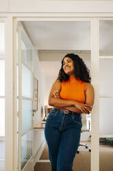 Cheerful female entrepreneur looking away with a smile while standing in an office with her arms crossed. Happy young businesswoman working in a creative workplace. - JLPSF07300